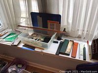 Wide view of drafting tools displayed on table by window, showing protractor, rulers, boxes, and bills box.