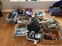 Several plastic bins and cardboard boxes containing various black, white, and orange cables and extension cords on a wooden floor