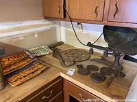 Photo showing the vintage weighing scale with bowl, metal weights, flywheel, and a few license plates on a wooden corner countertop with cabinets above.