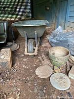 Photo shows a green wheelbarrow, round planter pot, and two round stepping stones on the ground in a partially covered outdoor area. Items appear weathered and used.