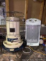 Front view of the two heaters side-by-side on basement floor, showing Kero Heat fuel heater with safety cage and Holmes electric heater with mesh front.