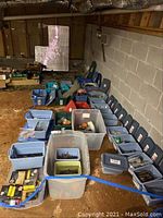 Long view showing multiple plastic storage bins of various sizes filled with assorted hardware components arranged on a concrete floor in a basement.