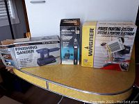Lot of three power tools in original boxes on table: Black & Decker finishing sander, rechargeable cordless screwdriver with charger, Wagner power painting system.