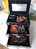 The open jewelry box showing various brooches, bracelets, pearl necklaces, and earrings inside the two lower drawers and ring slots on top.