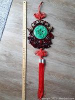 Full view of large Chinese hanging ornament laid next to a wooden ruler for scale, showing the top knot, jade green medallion with Chinese characters, surrounding reddish-brown carved resin, and long red tassel.