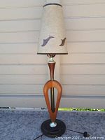 Full view of tall mid century table lamp showing fiberglass shade with two brown leaf-shaped designs, dark trim on top and bottom, wooden open frame stand, and black metal base.