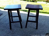 Pair of two wooden stools with dark finish shown outdoors on concrete driveway in sunlight.