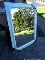 Full view of the large rectangular mirror leaning outdoors showing antique white ornate frame with corner protectors on all four corners.