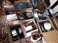 Eight boxes containing assorted kitchen cookware, appliances, bowls, and utensils on patterned carpet.