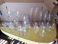 Various clear glass drinkware including multiple wine glasses, champagne flutes, and cocktail glasses set on yellow surface inside a cardboard box.
