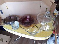 Full lot on yellow surface: includes brown Pyrex bowls, covered casserole, clear glass cake stand with dome lid, clear glass gravy boat, and small glass bowls.