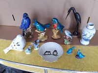Lot of vintage bird figurines, white dish, and blue and white Chinese porcelain bottle displayed on a yellow surface with cardboard background.
