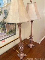 Pair of matching table lamps with brown metallic painted metal bases and beige silk fabric shades, shown on wooden surface with a painting in the background.
