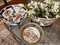 Three Asian decorative items displayed on a glass table over a patterned rug, including a large blue and white bowl, a smaller bowl with an Asian scene, and a large tureen with silk flowers.