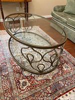 Side angle view of the oval glass top coffee table showing curved metal base and clear glass surface on patterned rug.