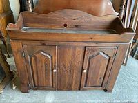 Front view of dry sink showing carved wheat pattern on backsplash and door panels, white porcelain knobs, dark stained wood finish.