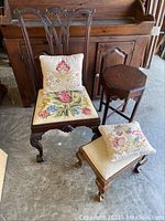 Photo showing Queen Anne style mahogany side chair with needlepoint upholstered seat, hexagonal side table, mahogany stool with cream seat, and two needlepoint embroidered pillows.