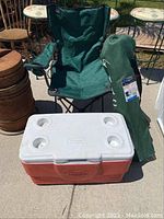 Two green MAC Sports Captain camp chairs folded, with one chair standing and the other in a carrying bag, alongside a red and white Coleman cooler with a white lid having four cup holders.
