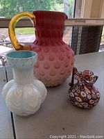 View of all three glass items including the large pink bobble pitcher, white geometric vase with blue interior, and small millefiori pitcher