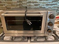 Full front view of the Cuisinart stainless steel toaster oven placed on a stove top, showing the closed door with the electrical cord hanging over the top and control knobs on the right.