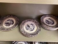 Set of dessert plates on shelf, showing stacks of plates with intricate scenic designs and patterned borders.