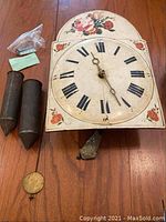 Front view of the clock face with two pine cone shaped weights, pendulum, and some accessories on wooden floor.