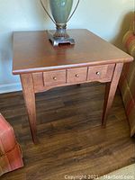 Wooden side table with drawer, placed on wooden floor next to armchairs, showing front and one side.
