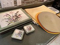 Photo showing stacked Pimpernel England rectangular floral placemats and square coasters with botanical illustrations nearby beige linen placemats and round hot plates/trivets.