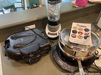 Shows three kitchen items on countertop: NuWave induction cooktop with pot and lid, Black & Decker blender, and black electric griddle.