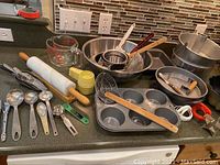 Wide view of kitchen counter with measuring spoons, rolling pin, mixing bowls, muffin tin, graters, and other baking and kitchen items.