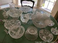 Wide view of various cut and pressed glassware items including bowls, cake stand, platters, and cruets arranged on table