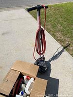 Full view of the Oreck floor cleaner standing upright on a driveway with long red power cord wrapped around handle, next to an open box with cleaning solutions.