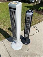 Two tower fans outdoors on concrete and grass. One white Holmes brand, one black with silver grill.