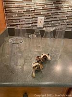 Overview of nine clear glass lamp chimneys arranged on countertop with three brass lamp burner assemblies and cotton wicks in front.