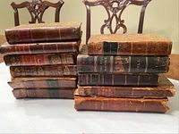 Photo showing a stack of 11 small antique books with worn leather and cloth bindings, titles faintly visible on the spine.