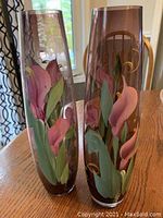 Two tall glass vases with hand painted pink and purple flowers and green leaves on purple tinted glass, displayed on wooden table near window.