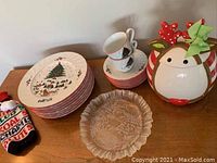 Christmas-themed dishes displayed together including plates, cups, saucers and ceramic plate with winter cabin scene, alongside reindeer cookie jar.