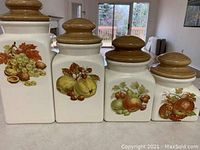 Front view of all four white ceramic food canisters with wooden lids, showing fruit designs on each.