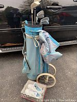Full view of light blue golf bag with five golf clubs and attached wheels, placed beside a car, outdoors.