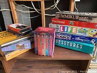 Image showing an assortment of board games stacked, including Rummikub, Parcheesi, The Game of Life SpongeBob SquarePants Edition, Sorry! Disney Edition, and Guesstures. Four purple boxed VHS tapes of The Wild Thornberrys main show volumes 1 to 4 on the left side of the table. Several boxed puzzles underneath the VHS tapes