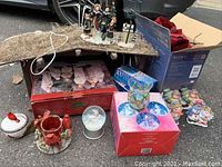 Wide view showing nativity set, tree skirt, plates, ceramic figurines, and other assorted Christmas decorations arranged on the ground near vehicle tire.