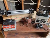 Four cameras and aluminum tripod holder arranged on a table, showing Pentax camera in box, Kodak camera and folded tripod.