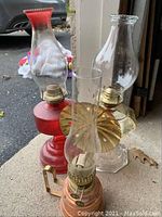 Three kerosene lamps positioned outdoors on a concrete surface showing varying base styles and glass chimneys.