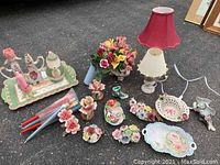 Wide view of all items included in the lot arranged on ground, showing various porcelain floral pieces, artificial flower arrangement, two lamps, and a tray with tea set.