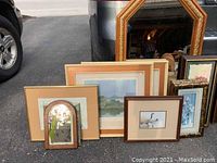 Multiple framed pictures including floral prints and a golden framed mirror reflect light in front of a parked vehicle.