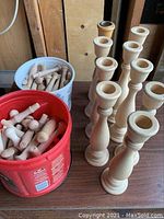 Nine unfinished wood candle holders standing upright next to two buckets of wooden pegs on a table.