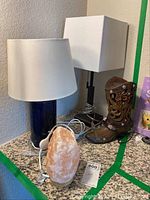 Four lamps on a corner table: black ceramic lamp with white tapered shade, black lamp with square white shade, cowboy boot lamp, and pink salt lamp with cords visible.