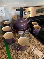 Photo showing six purple Pfaltzgraff mugs surrounding matching plates on kitchen counter near stove.
