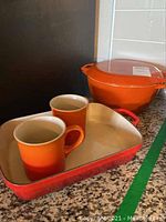 Two stoneware mugs inside a rectangular stoneware tray and a covered round cast iron crock in the background, all in Le Creuset's signature colors.