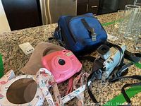 Image showing both the pink Fujifilm Instax Mini instant camera with floral strap and Canon handheld video camera with strap alongside their cases on a kitchen countertop.